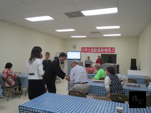 高建智由莊雅淑陪同參觀歡樂歌唱班耆老練唱情形。與歡樂歌唱班會員鐵大鵬握手問候，會長陳德新(著紅色T恤衫者)表示歡迎